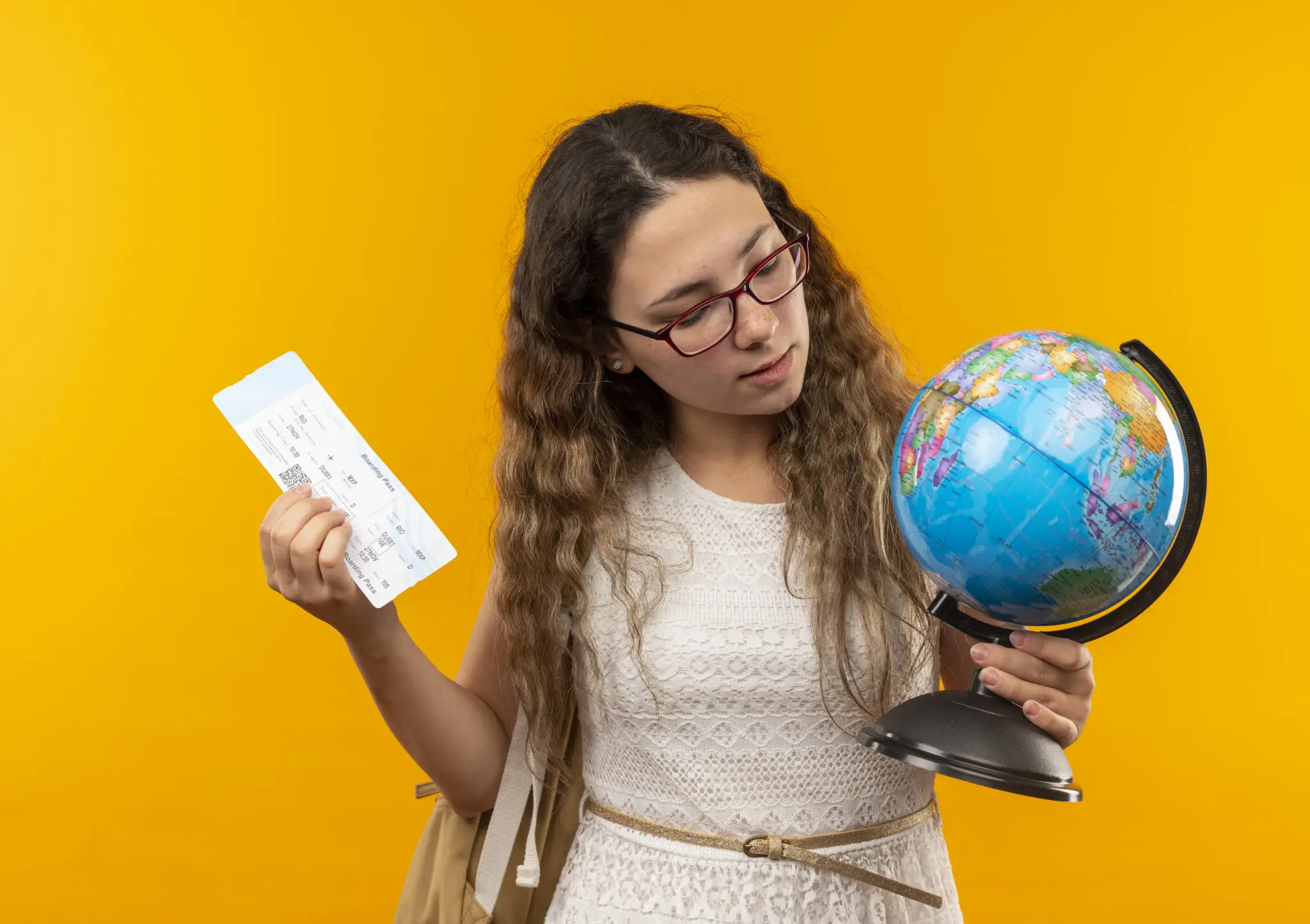 confident young pretty schoolgirl wearing glasses and back bag holding airplane ticket and globe looking at globe isolated on yellow background etias visto europeu