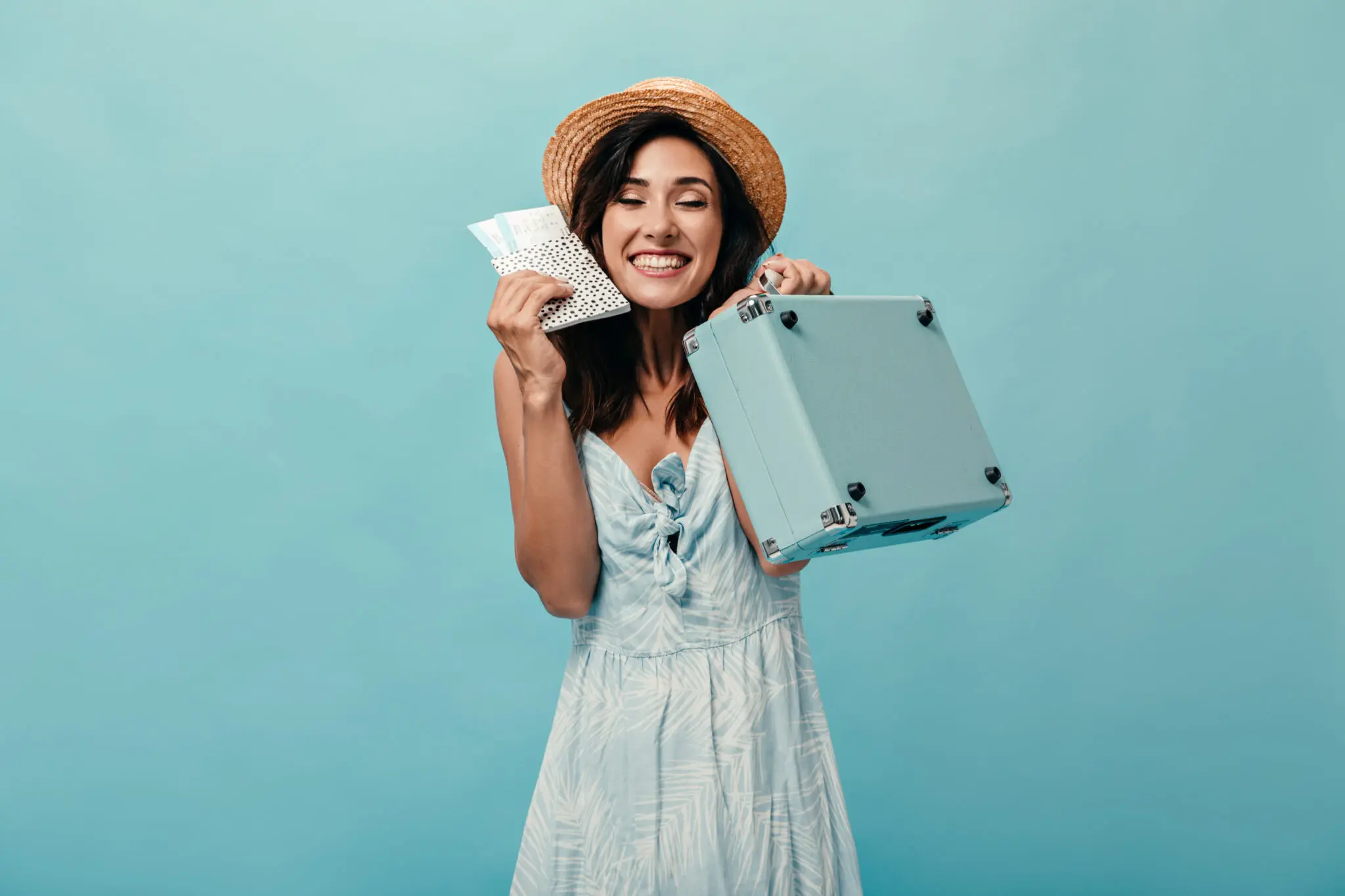 Girl in joyful mood holds vacation tickets and blue suitcase. Woman in straw hat with smile on her face posing on isolated background. Real Seguro Viagem